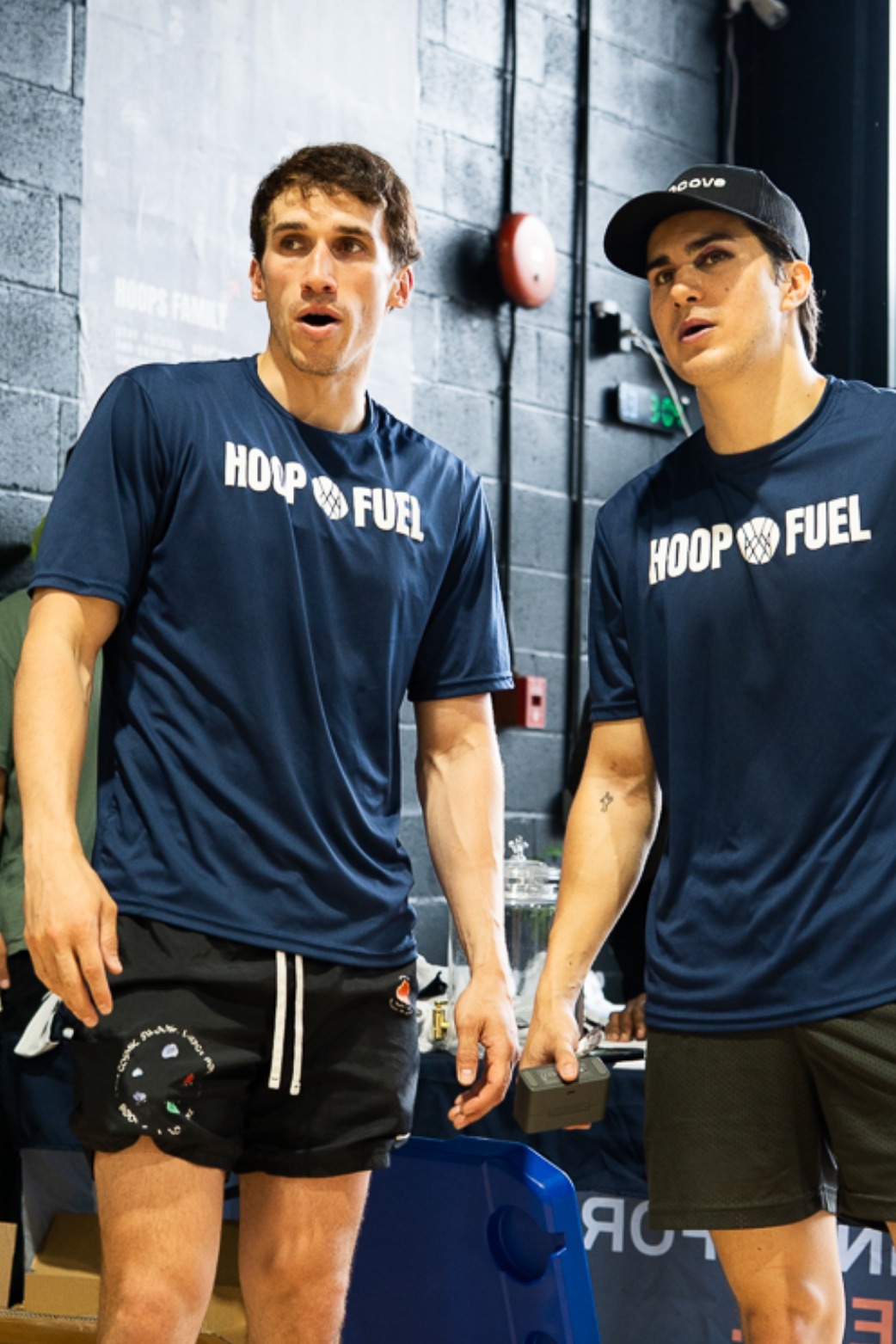 Two men wearing 'HOOPFUEL' t-shirts in a gym setting.